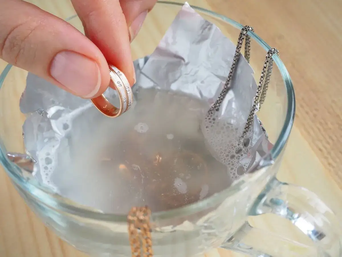 Mano sosteniendo un anillo de oro y plata, sumergiéndolo en agua con burbujas para limpiar joyas. Una cadena plateada cuelga del borde del vaso.