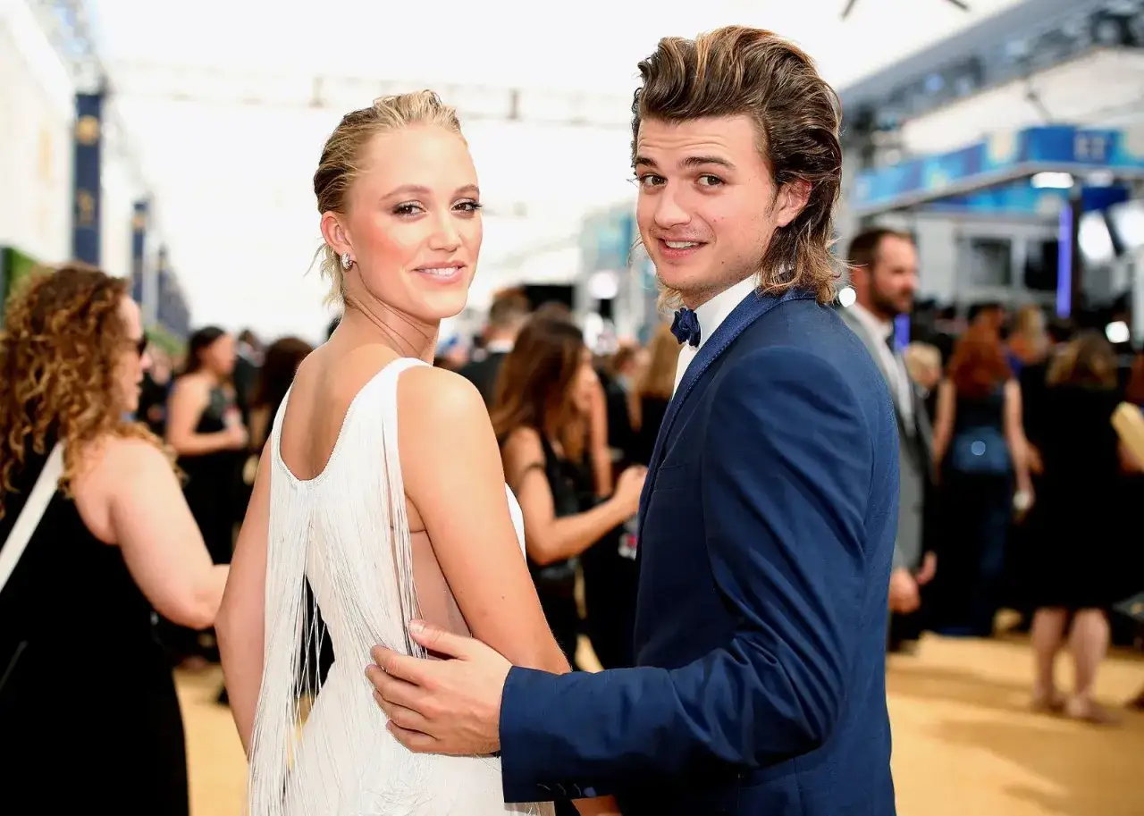 Joe Keery y su pareja, ambos sonriendo a la c&aacute;mara en una alfombra roja. Ella lleva un vestido blanco con flecos, &eacute;l un traje azul.