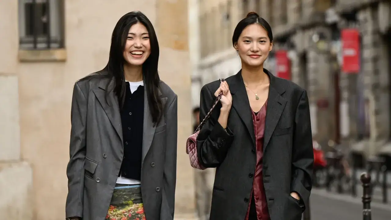 Dos mujeres lucen un elegante outfit oficina: una con blazer gris y falda floral, la otra con blazer oscuro y top burdeos.
