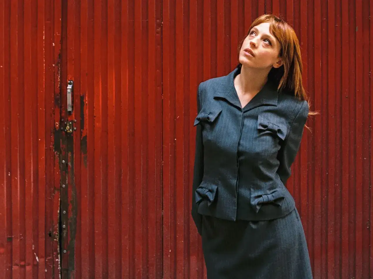 Catalina Sopelana, con su traje gris y cabello rojizo, mira hacia arriba contra una pared roja acanalada.