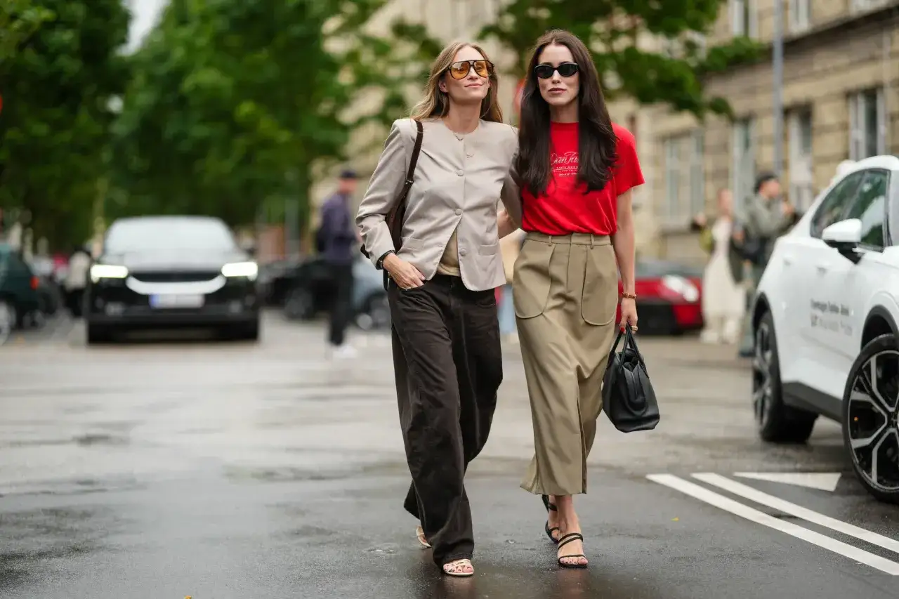 Dos it girls pasean con estilo por la calle. Una lleva un blazer beige y pantalones anchos marrones, la otra un top rojo y falda beige.