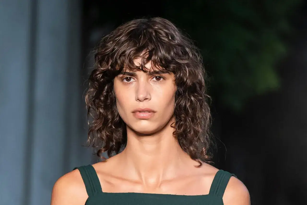 Mujer con cabello rizado y oscuro, con un estilo de flequillo cortina que enmarca su rostro.