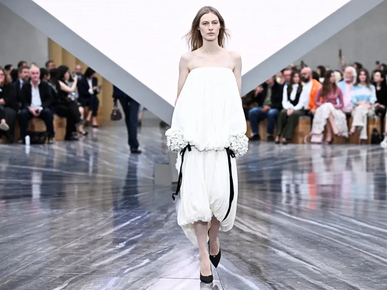 Modelo desfila un vestido blanco voluminoso con detalles florales y cintas negras, una creaci&oacute;n de Jonathan Anderson, ante un p&uacute;blico atento.