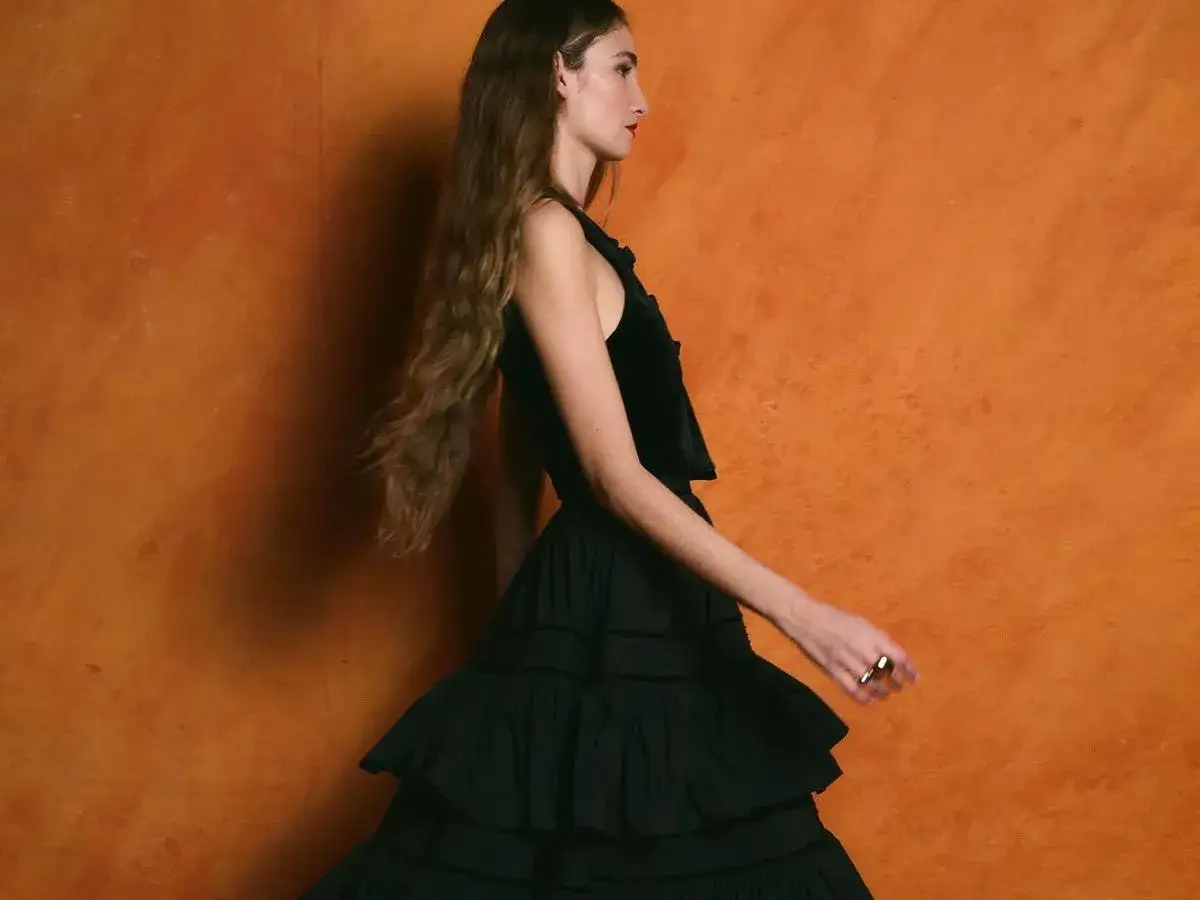 Almudena Cañedo, con su melena suelta y un vestido negro voluminoso, posa con elegancia ante un fondo naranja vibrante.
