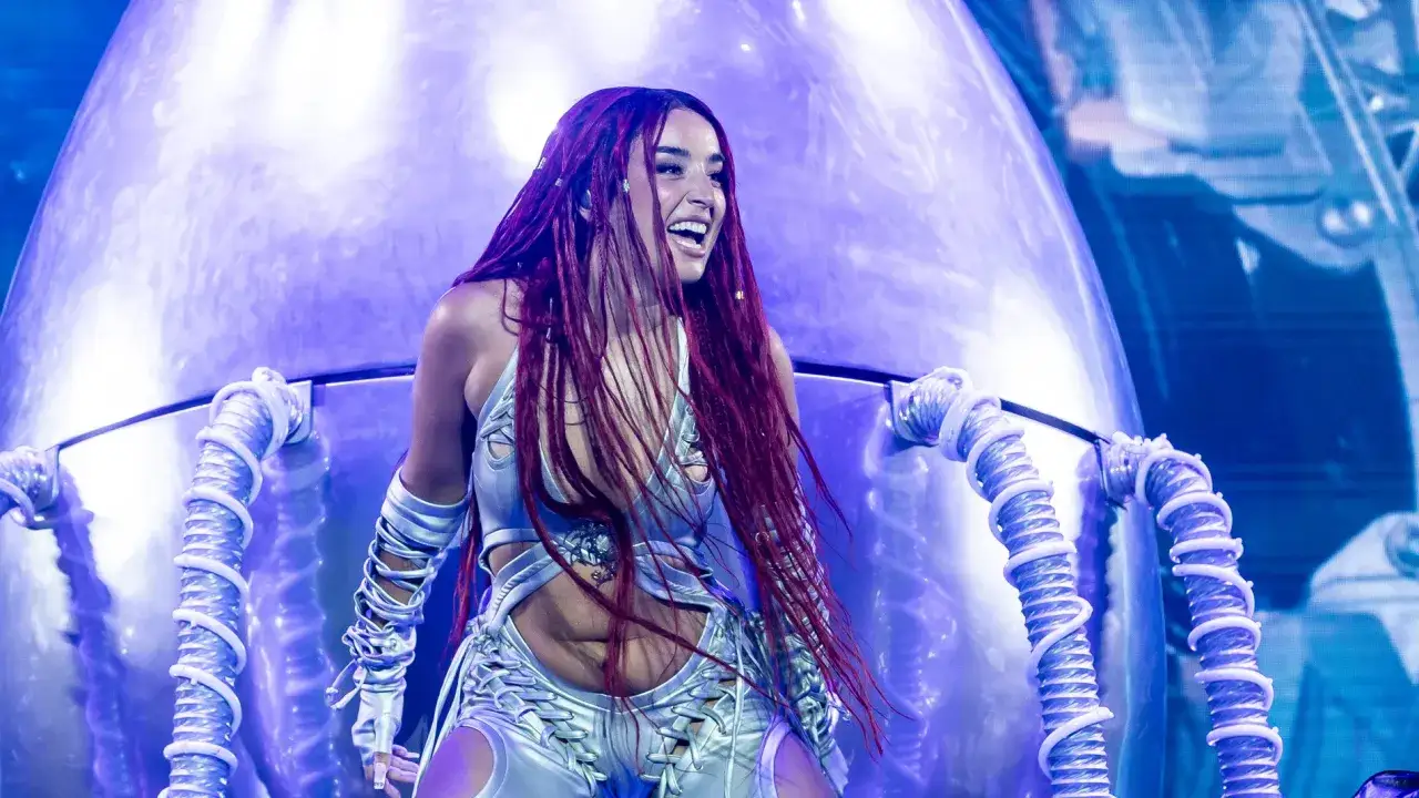 Lola Indigo, con su cabello largo y rojizo, sonríe en un escenario futurista, vestida con un traje plateado brillante.