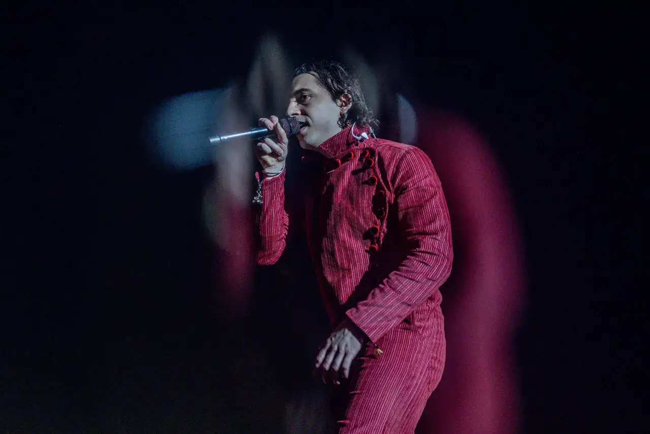 Ralphie Choo canta en un escenario con un traje rojo a rayas, iluminado por luces tenues.