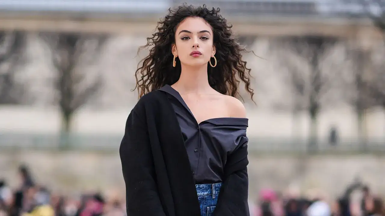 Deva Cassel, con su cabello rizado y mirada intensa, luce un estilo audaz con una camisa desabrochada y un abrigo negro.