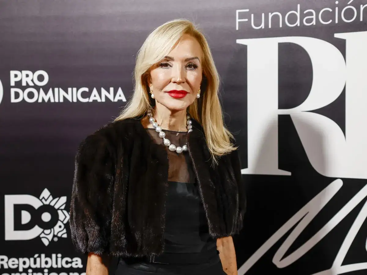 Carmen Lomana, elegante con abrigo de pelo y perlas, posa ante un fondo con el logo 