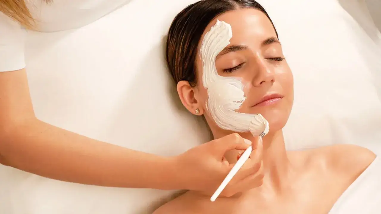 Una cosmetóloga aplica una mascarilla cremosa en el rostro de una mujer como parte de un tratamiento facial relajante.