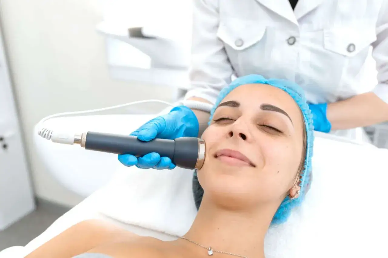 Una mujer disfruta de un tratamiento de radiofrecuencia facial. Un profesional con guantes azules aplica el dispositivo en su mejilla.