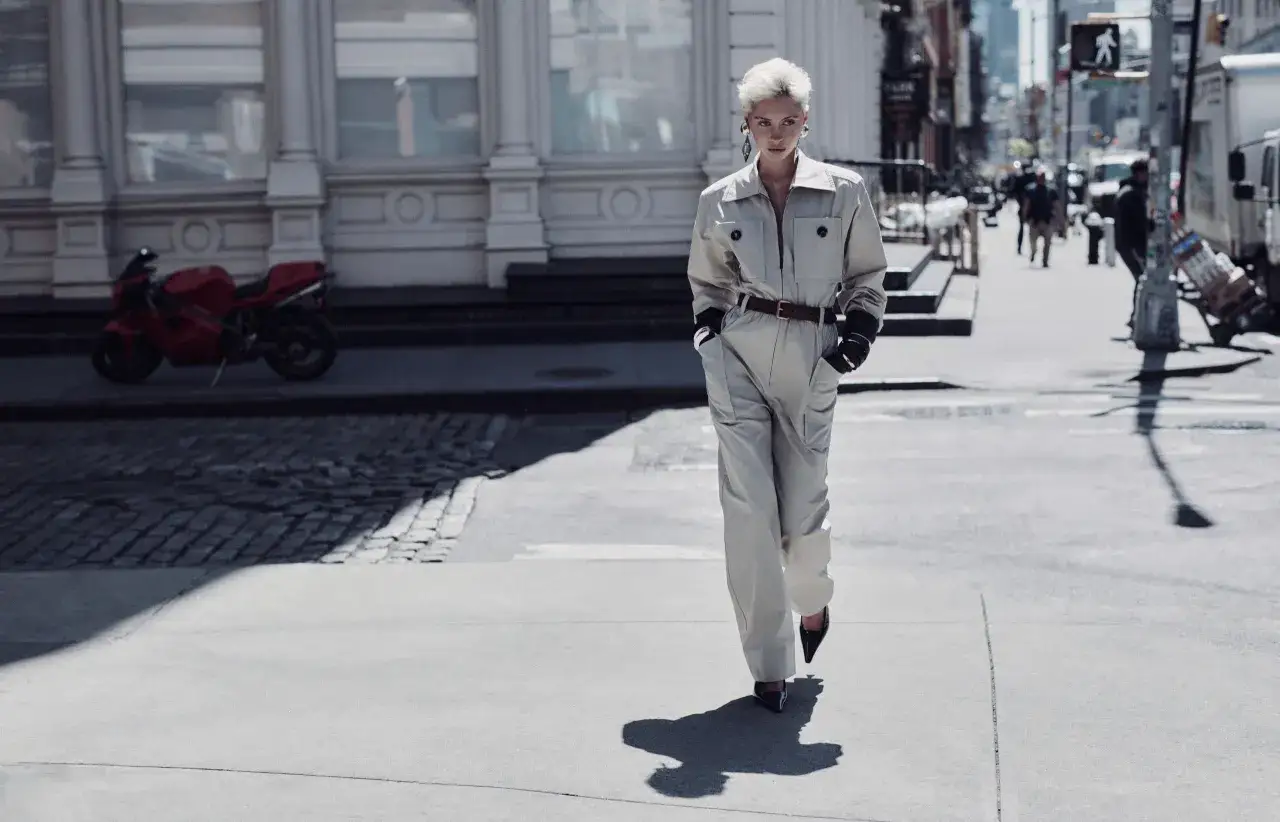 Iris Law camina por la calle con un mono beige, guantes negros y tacones. Una moto roja aparcada al fondo.