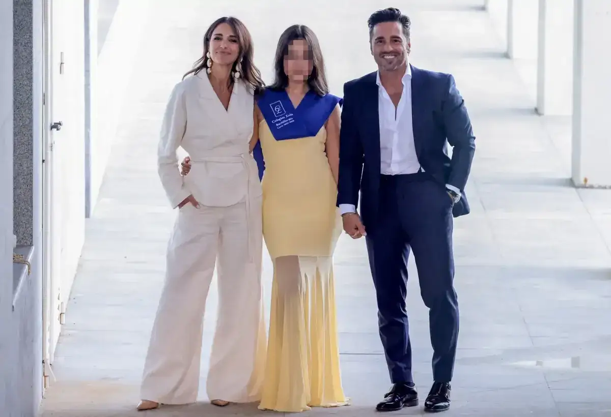 Familia celebra la graduación de su hija. La madre, vestida de blanco, abraza a la graduada con su toga azul. El padre, de traje azul, sonríe orgulloso.