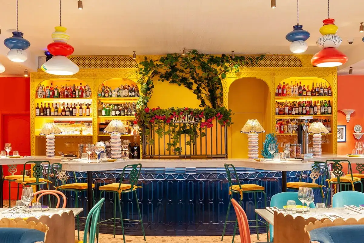 Bar vibrante con estanterías amarillas llenas de botellas, lámparas de mimbre y sillas coloridas. Un lugar perfecto para disfrutar de una beata pasta.