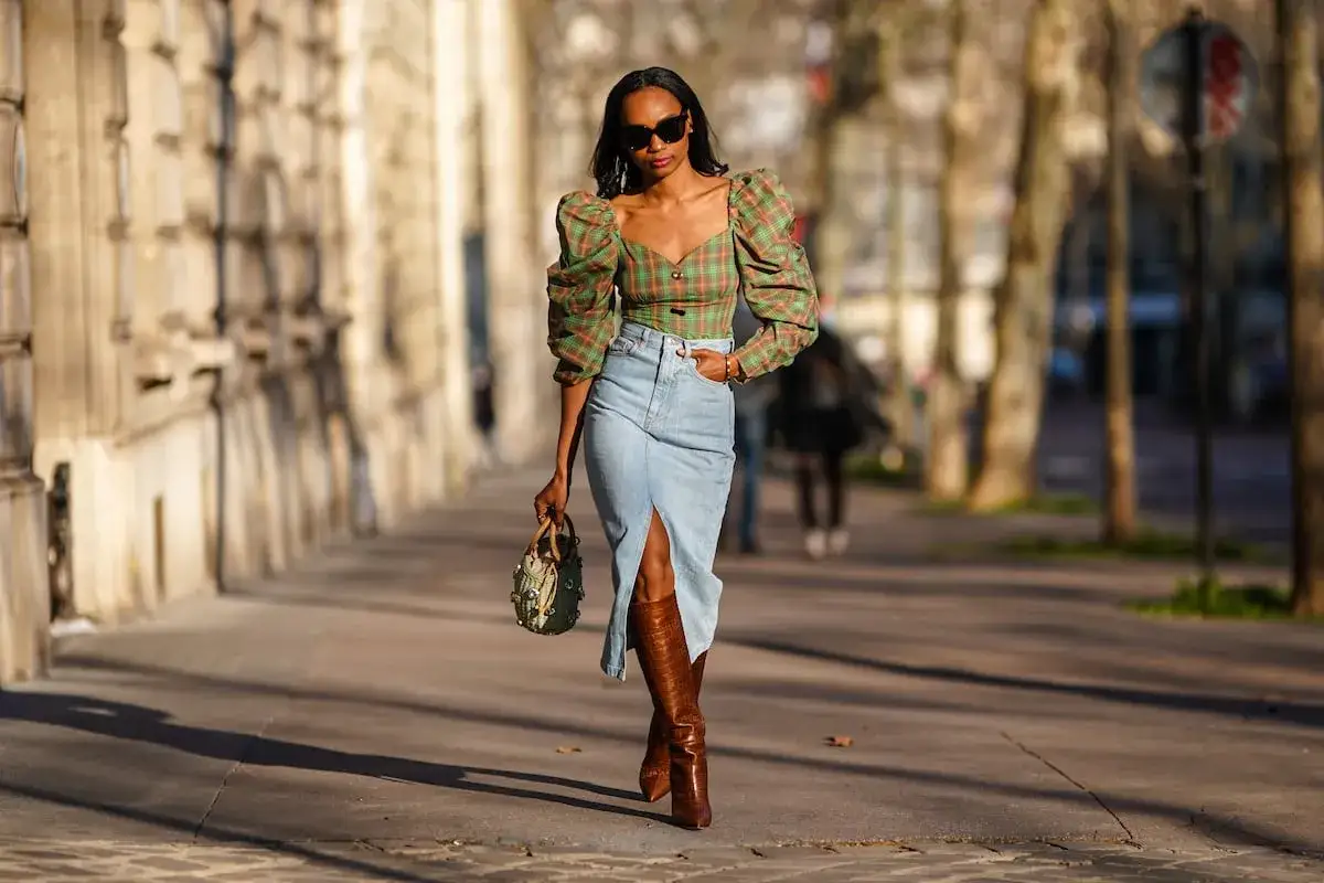 Mujer con top de cuadros y falda vaquera larga con abertura, botas altas y bolso.