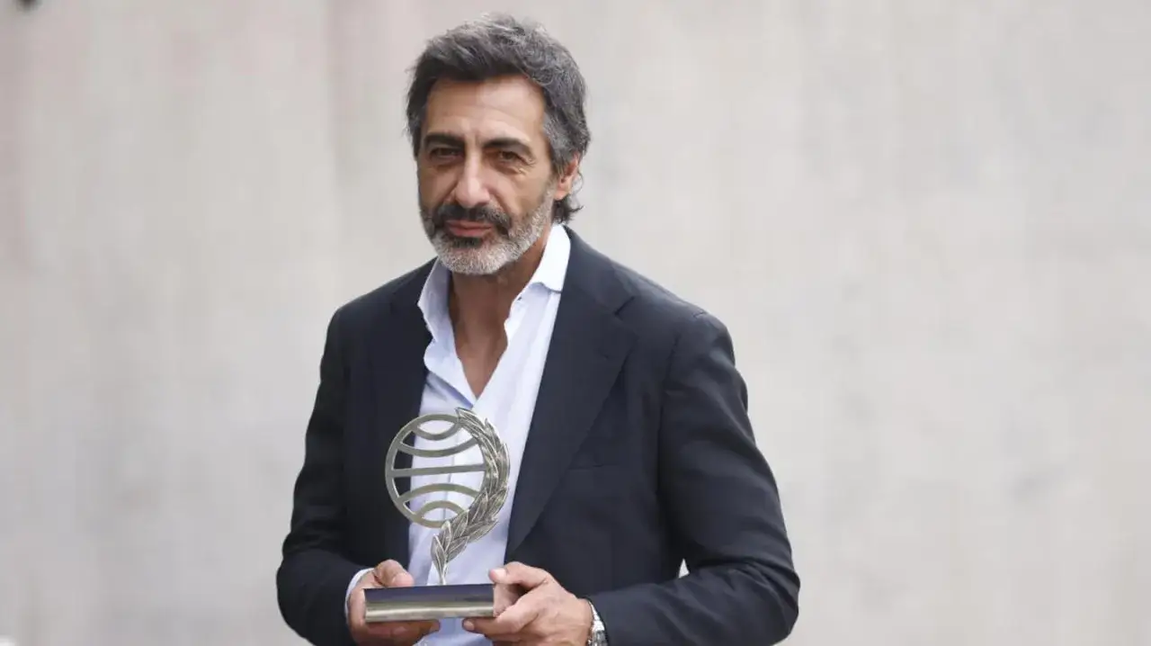 Juan del Val, con barba canosa, viste traje oscuro y camisa azul clara, sosteniendo un trofeo con un globo terráqueo y laureles.