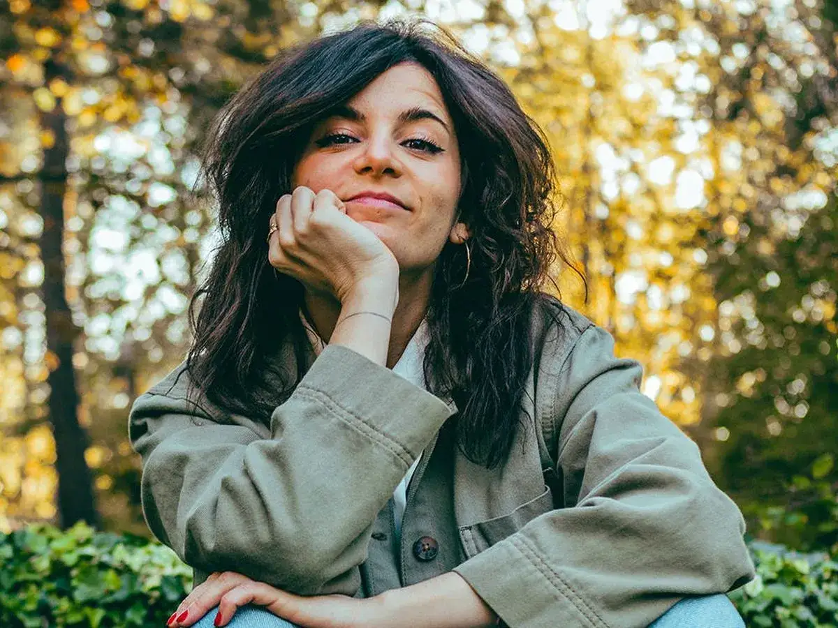 Ana Iris Simón, con su mano apoyada en la barbilla, sonríe pensativa entre árboles otoñales.
