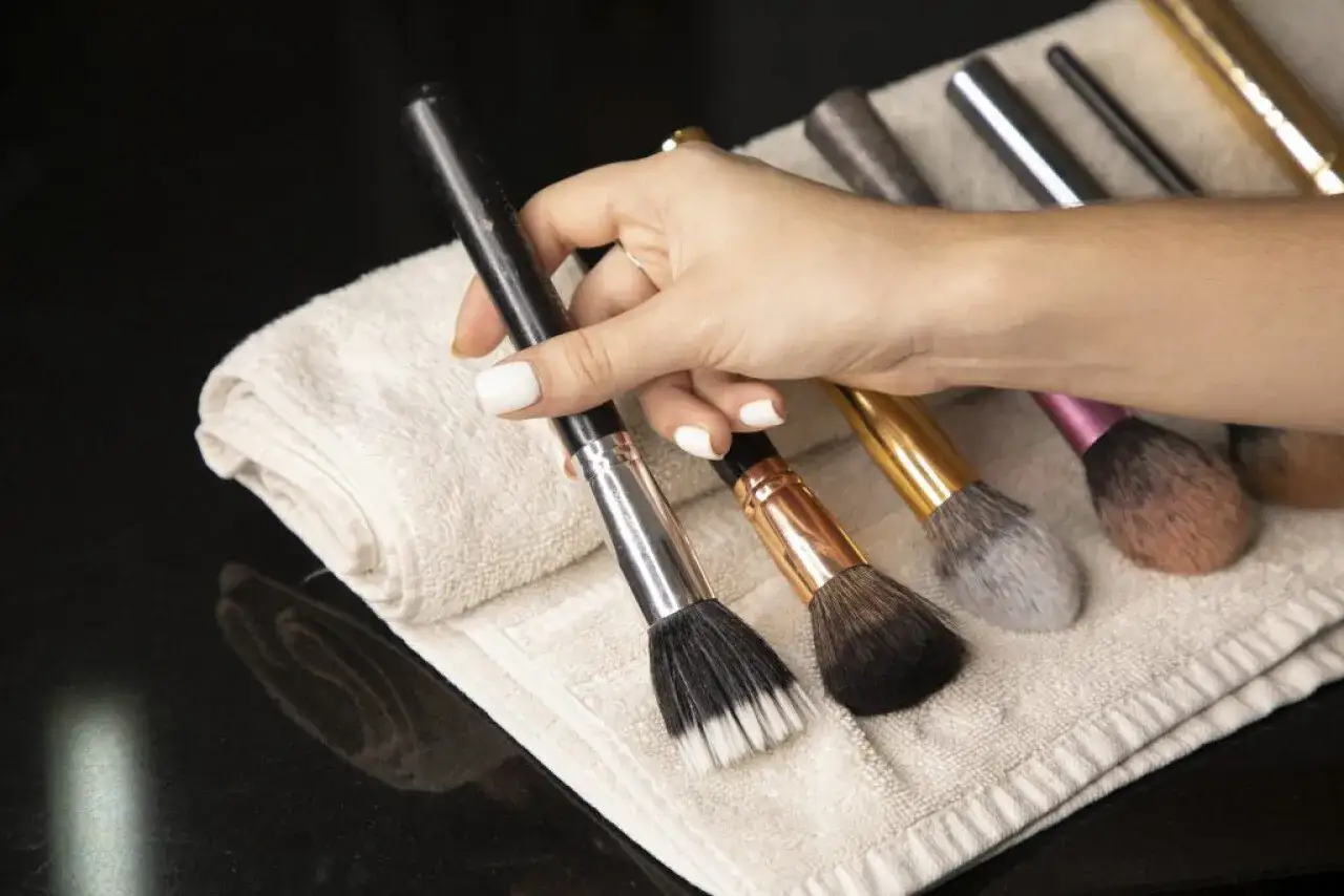 Mano con uñas blancas sosteniendo un pincel de maquillaje, junto a otros pinceles sobre una toalla. Es hora de limpiar brochas de maquillaje.