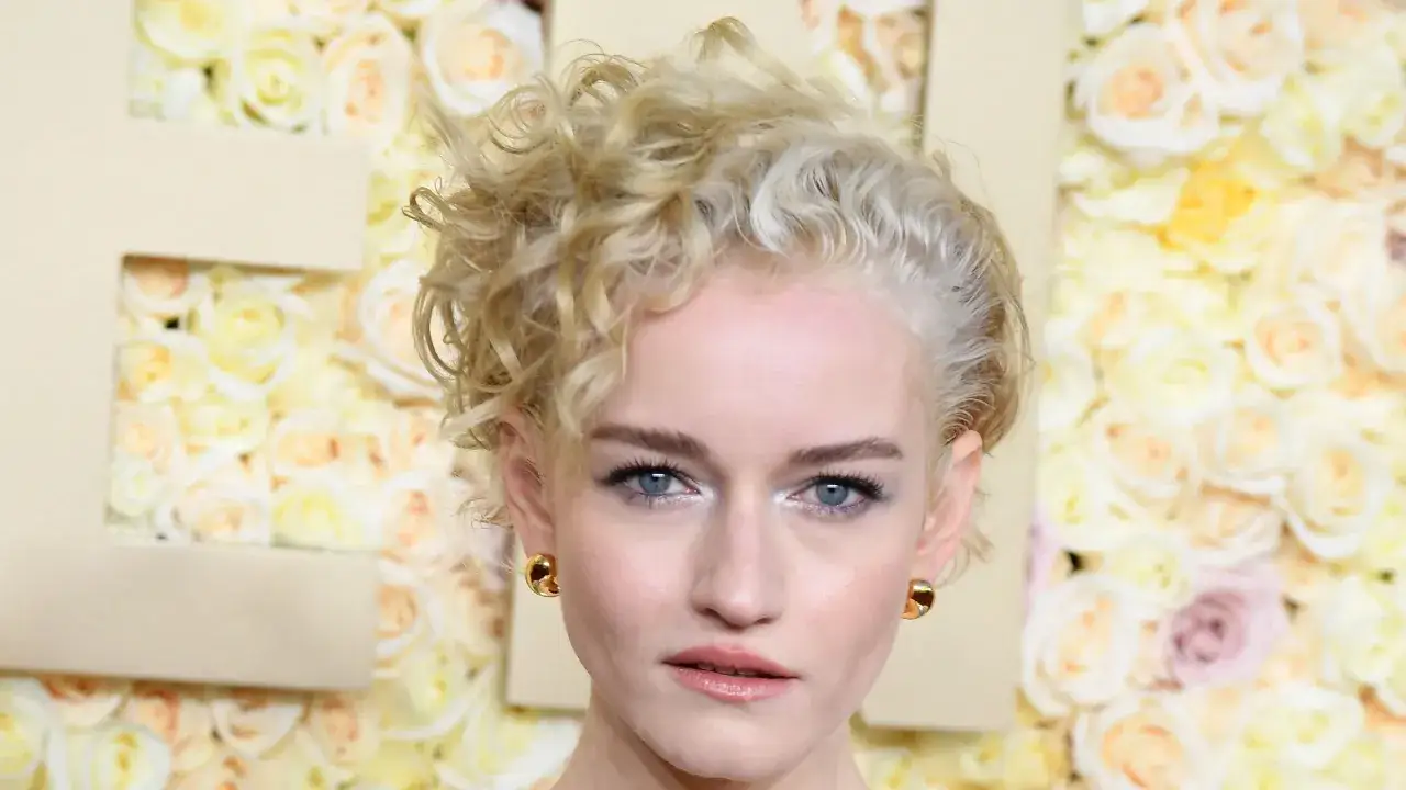 Mujer con pelo corto rizado rubio, ojos azules y pendientes dorados, posando ante un fondo de rosas.