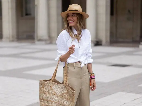 Mujer con sombrero de paja, camisa blanca anudada y vaqueros beige. Lleva un bolso de mimbre y pulseras coloridas.