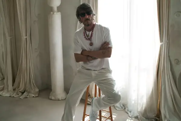 Pierpaolo Piccioli, con gafas de sol y collares llamativos, posa con los brazos cruzados en un entorno elegante con cortinas y columnas.