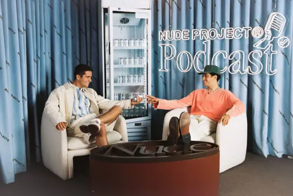 Los fundadores de Nude Project brindan con latas de su bebida, sentados en sillones frente a un cartel de neón del podcast de la marca.