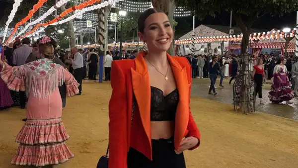 Chica sonriente con chaqueta naranja y top negro, lista para la ropa para ir a la feria de dia. Ambiente festivo con luces y gente.