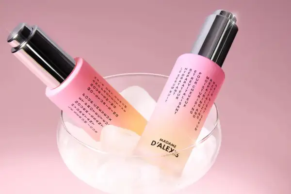 Dos frascos de aceite para pelo, con un degradado rosa y amarillo, reposan sobre cubitos de hielo en una copa.