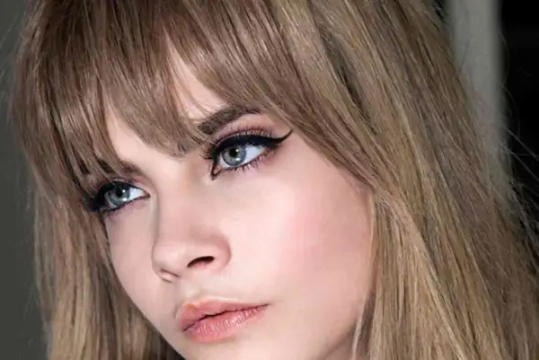 Primer plano de una mujer con flequillo rubio desfilado, delineado de ojos dramático y labios rosados.