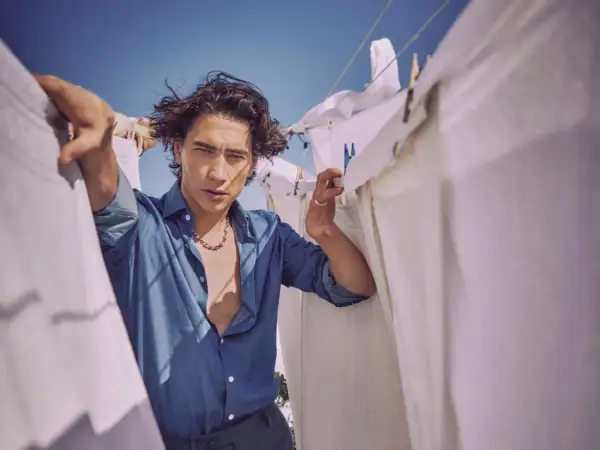 Enzo Vogrincic, con su camisa desabrochada y collar llamativo, posa entre sábanas blancas tendidas al sol.