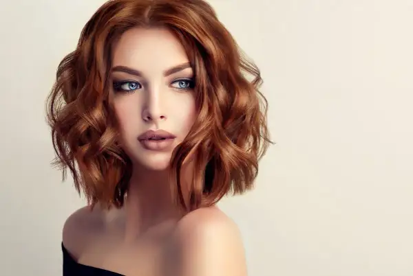 Mujer con cabello cobrizo ondulado en un elegante corte midi. Sus ojos azules y labios definidos resaltan su belleza.