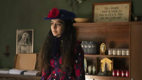 Irene Balmes, con un sombrero azul y un vestido floral, mira pensativa. Detrás, estanterías con objetos religiosos y un cuadro.