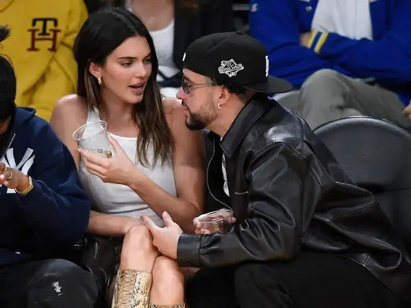 Bad Bunny y Kendall Jenner conversan en un evento deportivo, ella con un vaso en la mano y él con chaqueta de cuero.
