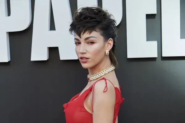 Mujer con un llamativo mullet corto, maquillaje rojo y vestido rojo, luciendo un collar de perlas.