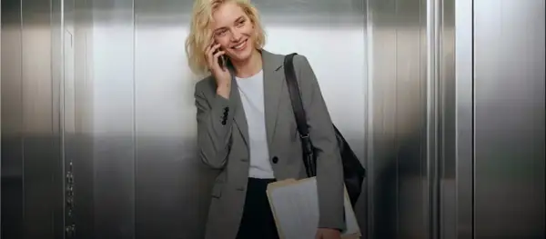 Mujer sonriente en ascensor, con un elegante outfit oficina, hablando por teléfono y cargando carpetas.