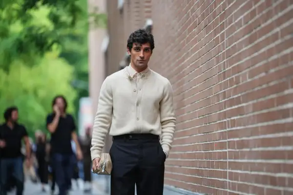 Hombre con suéter crema y pantalón negro, bolso de mano beige. Un ejemplo de las últimas tendencias moda hombre.